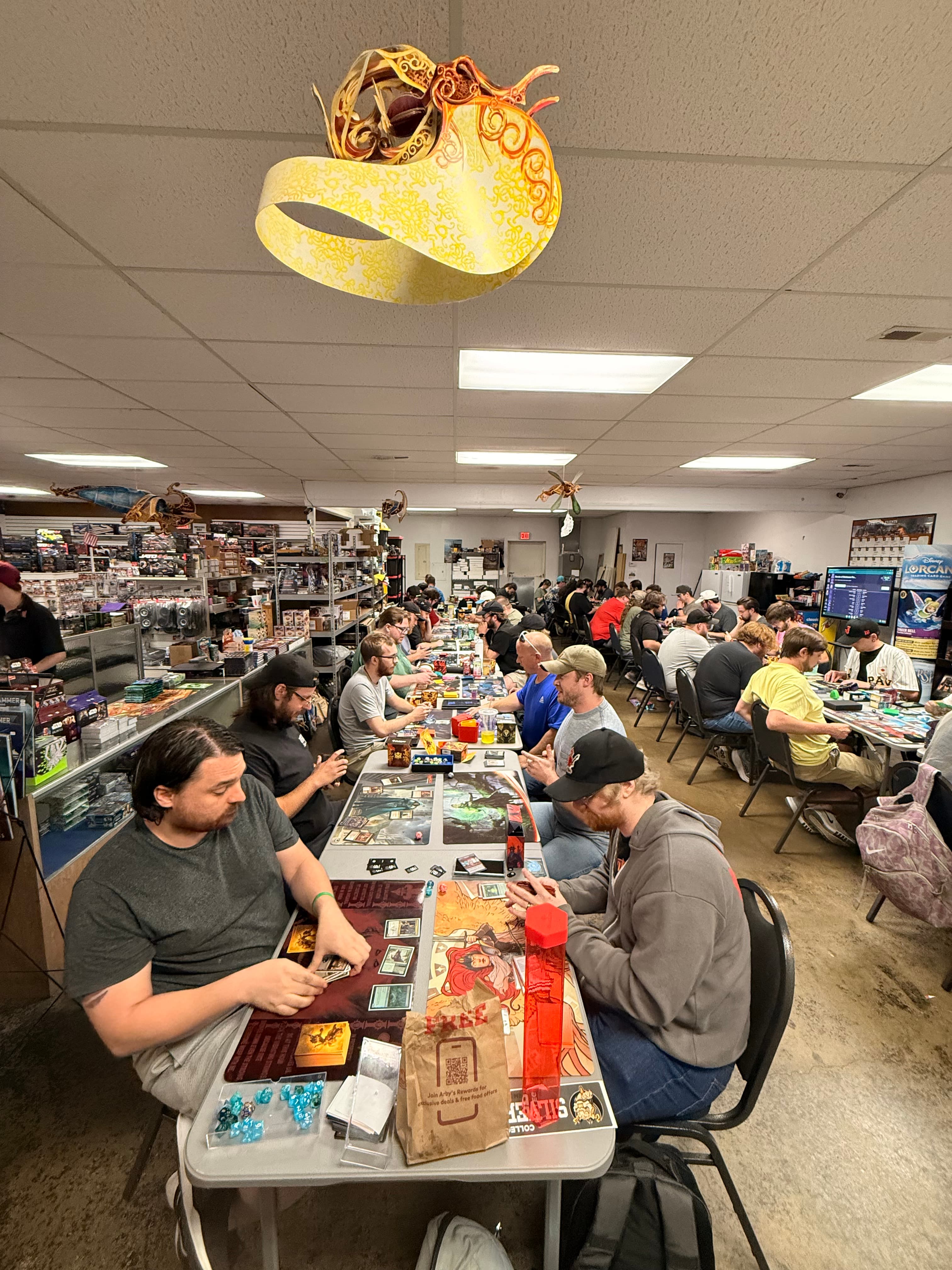 Gamers playing trading card games at long tables inside a crowded, brightly lit hobby shop.