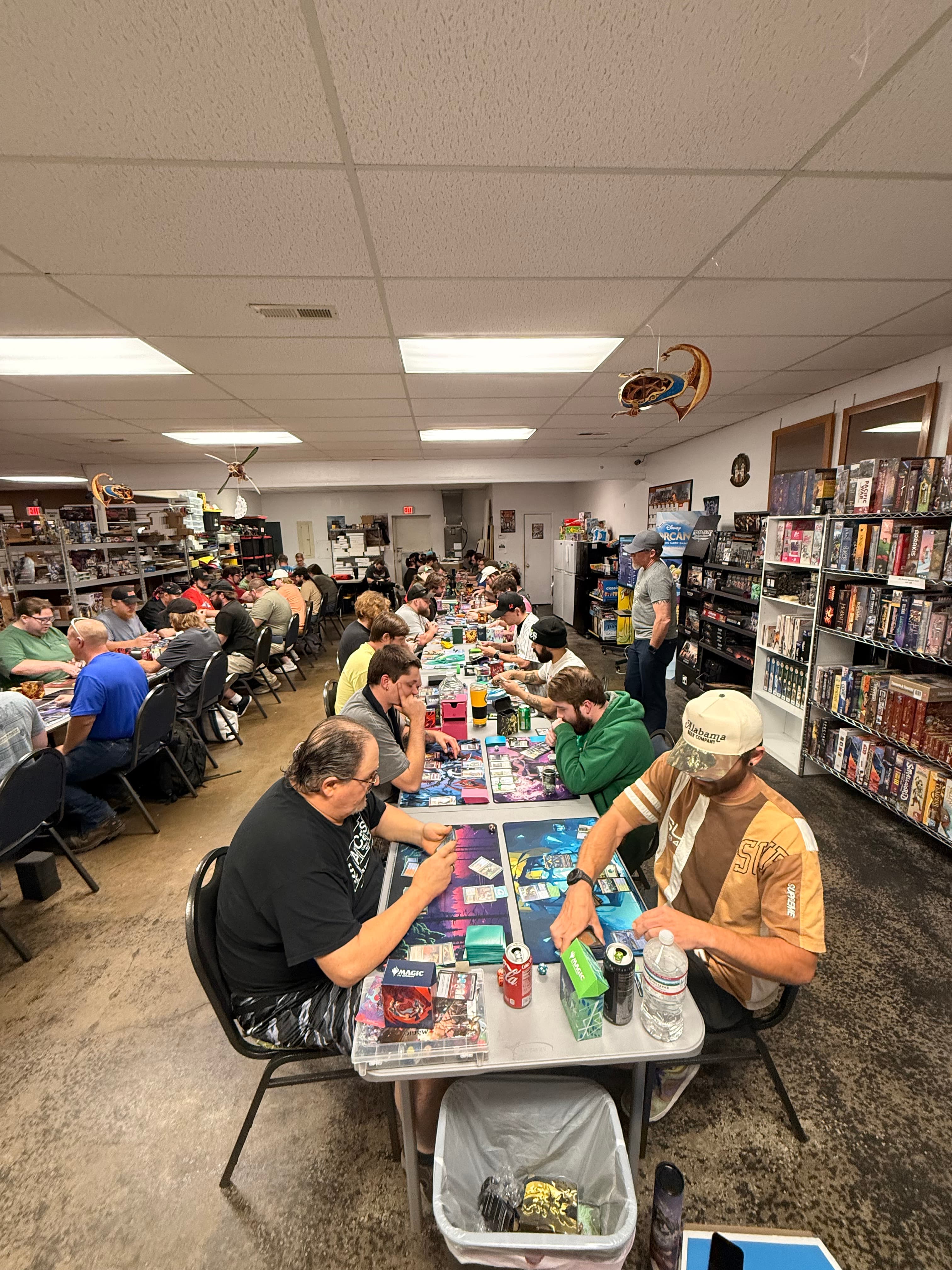 People seated at long tables in a crowded game store playing trading card games.