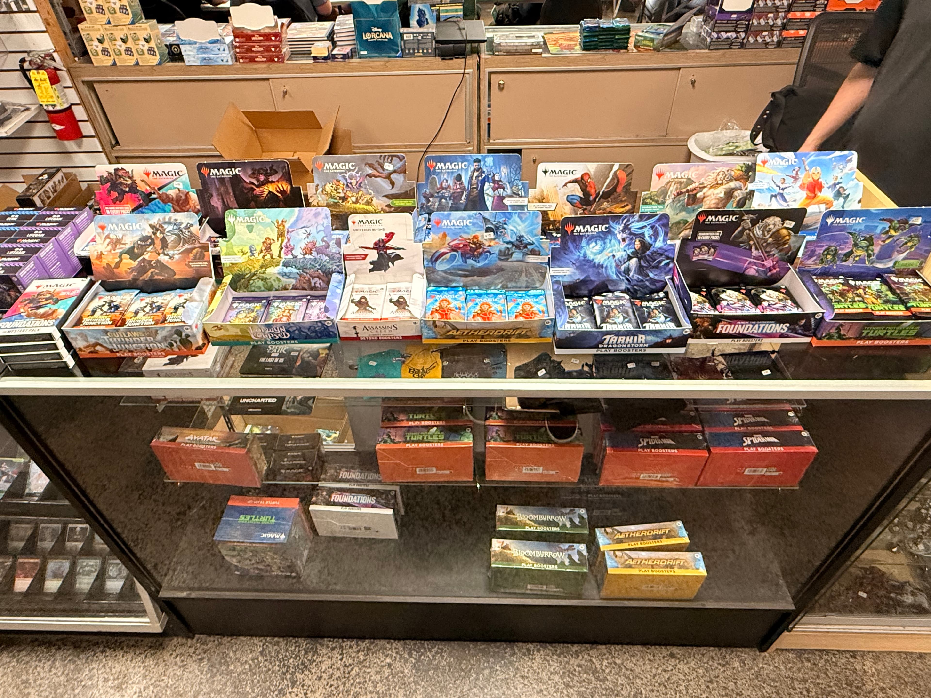 Glass display case showcasing various themed Magic: The Gathering booster boxes and card packs.
