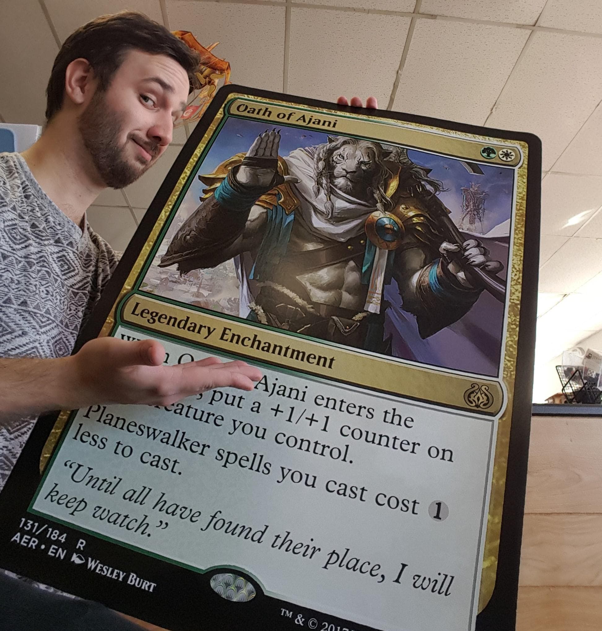 A man proudly displays an oversized Magic: The Gathering card titled 'Oath of Ajani'.